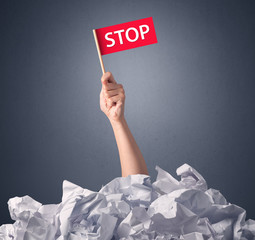 Female hand emerging from crumpled paper pile holding a red flag with stop written on it 