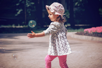 Naklejka premium A little Caucasian-looking girl in a panama dress plays with air-breathing bubbles on the nature, day, close-up