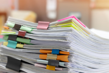 Pile of unfinished homework assignment and report separated by colorful papers and paper clips on teacher's desk waiting to be managed and checked. Stack of paperwork. Education and business concept.