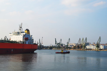 Fototapeta premium A small pilot ship leads a large red cargo ship at the seaport on a warm windless autumn day