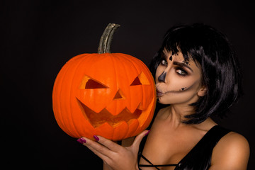 Sexy brunette woman in Halloween makeup and lingerie hold a pumpkin on a black background in the...