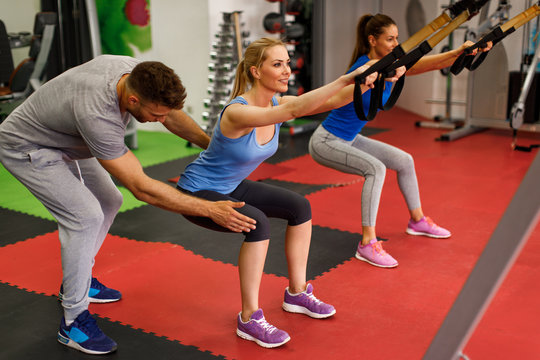 Personal trainer assisting young woman while exercising with trx in the gym