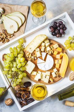Different types of cheese on wooden board, olive, fruits, almond and wine glasses on white tray