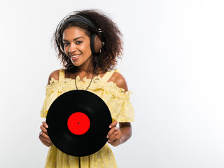 Young beautiful african american woman with headphones in yellow dress enjoying and dancing with vinyl record at white background