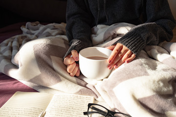 Having coffee and reading in bed on lazy sunny morning. Female hands holding steaming cup of hot...