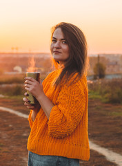 Young woman  in a orange sweater with thermos thermo cup outdoor portrait in soft sunny daylight. Autumn. Sunset. Cozy