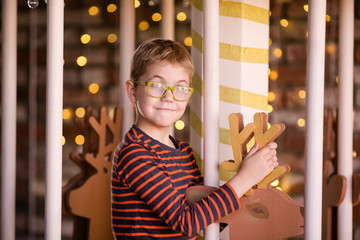 Fototapeta na wymiar Nice blonde boy with glasses on the new year carousel with wooden deer and bright lights