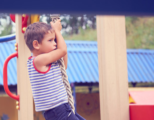 Little Caucasian boy in striped t-shirt hang the rope by his hand to exercise at out door playground in summer day.