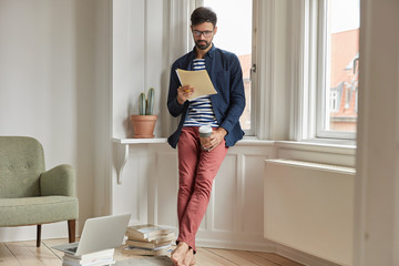 Full length shot of serious unshaven man or entrepreneur studies invoice, does paper work at home, holds takeaway coffee, stands near window sill, dressed in fashionable clothes, laptop computer near