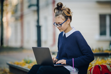 Close up Pretty hipster teen girl sitting on a sidewalk on autumn city street and working laptop computer. Schoolgirl using notebook outdoor. Beautiful autumn weather.