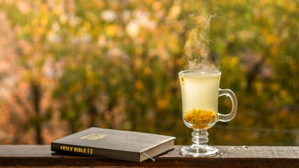 Cozy still life: cup of hot sea buckthorn tea and book Bible on vintage windowsill against warm...