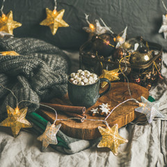 Christmas or New Year winter hot chocolate with marshmallows, cookies and cinnamon sticks over wooden board served with holiday light garland and grey sweater, selective focus, copy space, square crop