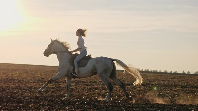 Young blonde girl riding on a horse on the field during sunset