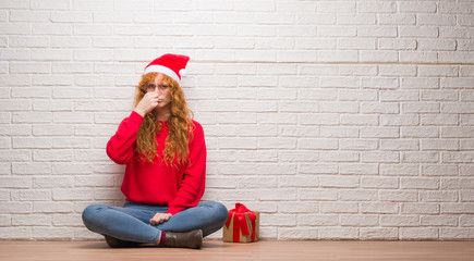 Young redhead woman sitting over brick wall wearing christmas hat smelling something stinky and disgusting, intolerable smell, holding breath with fingers on nose. Bad smells concept.