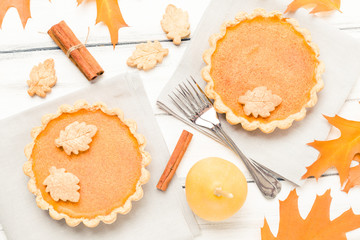 Pumpkin pie with cinnamon and cookies on gray napkins on white wooden background with autumn yellow leaves.