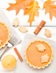 Pumpkin pie with cinnamon and cookies on gray napkins on white wooden background with autumn yellow leaves.