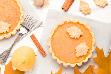 Pumpkin pie with cinnamon and cookies on gray napkins on white wooden background with autumn yellow leaves.