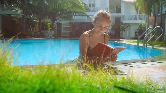 blonde girl in bikini reads book in pool leaning on barrier