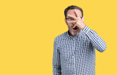 Handsome middle age elegant senior business man wearing glasses over isolated background peeking in shock covering face and eyes with hand, looking through fingers with embarrassed expression.