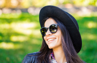 Portrait of happy mixed race young woman in hat. Hispanic hipster girl in the park. Lady wearing plaid shirt and sunglasses, trendy look.