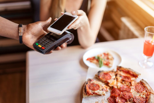 Blonde Female Customer Paying With NFC Technology On Smart Phone For Her Dinner.