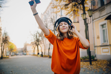 cute young girl listening music in headphones, dansing and holding mobilephone in hand, urban style, stylish hipster teen in black hat listen music on autumn city street, orange crazy street style