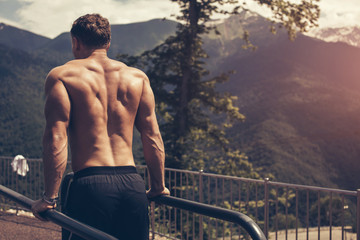 Backside view of muscular fair-haired european man in black shorts and naked top doing dips on parallel bars . Outdoor workout with blue sky and green mountains on the background and panoramic views. - Powered by Adobe