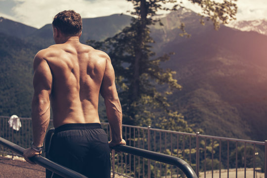 Backside view of muscular fair-haired european man in black shorts and naked top doing dips on parallel bars . Outdoor workout with blue sky and green mountains on the background and panoramic views.