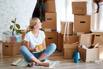 smiling woman with tea in hand sitting on floor of new apartment, pile of moving boxes on background