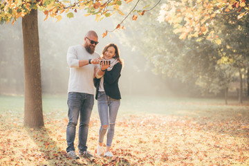 Young happy couple walking and having fun in autumn park and looking smartphone together