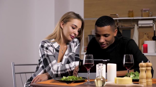 Happy smiling and laughing interracial couple looking at digital tablet PC while having candle light dinner in their kitchen. Dolly slider 4K footage