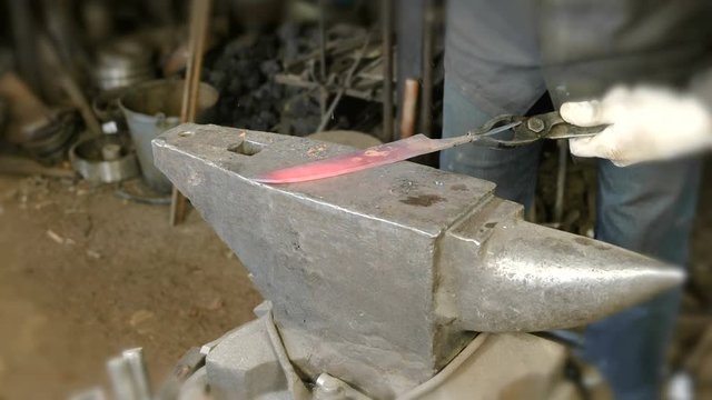 Making the knife out of metal at the forge. Close up blacksmith's hands hitting hot metal with a massive hammer on an anvil.