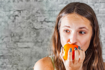 portrait of young teenager brunette girl with long hair eating persimmon kaki fruit on gray wall background