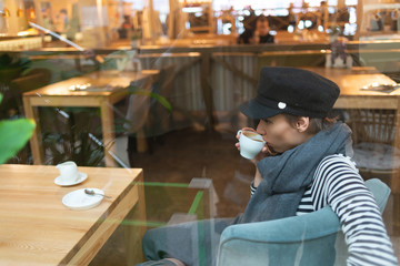 beautiful woman in hat and scarf drinking coffee while sitting in cafe
