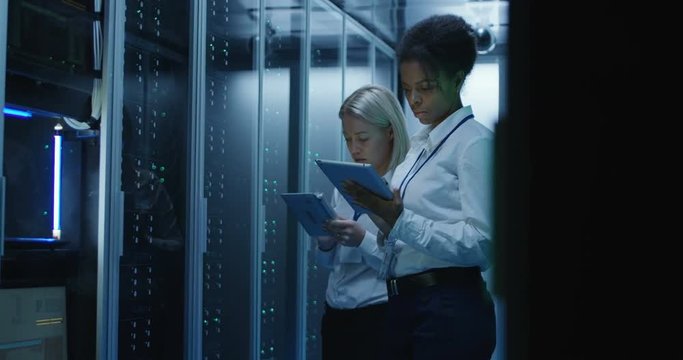 Medium shot of two women working in a data center with rows of server racks and checking the equipment and discussing their work