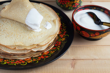 Fried pancakes, sour cream and a mug of milk in a wooden bowl with a bright Russian pattern on a wooden table