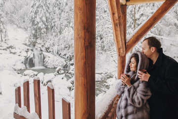 Stylish couple holding hot tea in cups and looking at winter snowy mountains from wooden porch. Happy romantic family with drinks hugging. Space for text. Holiday getaway together.