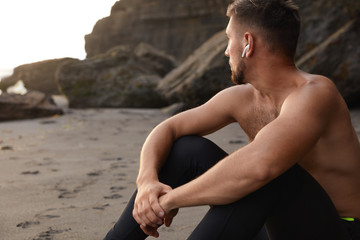 Horizontal view of contemplative sportsman sits on sand, focused aside into distance, poses half naked, enjoys calm atmosphere near ocean, listens pleasant music, goes in for sport regularly