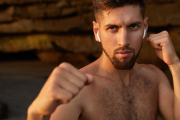 Obraz premium Strong angry boxer poses in defense gesture, clenches fists, has outdoor exercise, prepares for competitions, nas naked body, listens favourite music or radio with earphones. People and boxing concept
