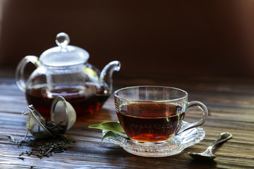 Cup of black tea, tea leaves, teapot, tea ball on wooden background.