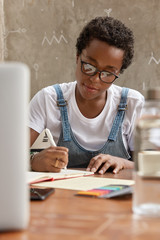 Boyish woman with short hairstyle, black skin, takes notes indoors, uses technology for work, holds...