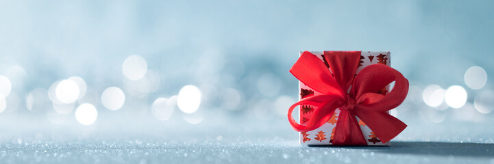 Beautiful red christmas gift with large bow on shiny blue background and defocused christmas lights in the background. Christmas banner with copy space.