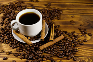 Cup of coffee, roasted coffee beans and cinnamon sticks on wooden table