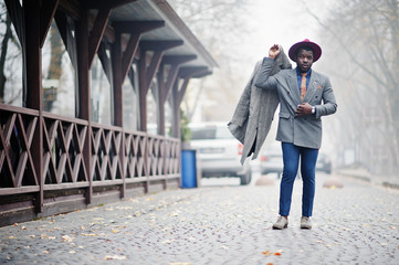 Stylish African American man model in gray coat, jacket tie and red hat posed at foggy weather street.