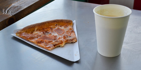 Bitten slice of pizza on a paper plate in a cafe and a paper cup of coffee