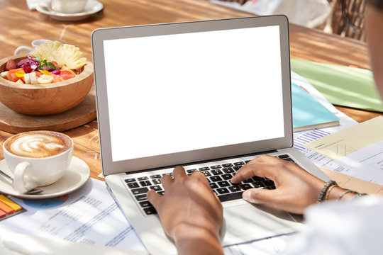 Unrecognizable young woman keyboards on laptop computer, mock up space for your advertising content, sits at table, enjoys coffee and fruit salad in cozy place, connected to wireless internet