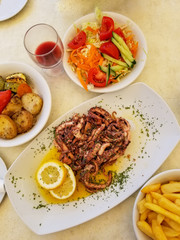 Fried octopus with lemon and spices, vegetables and french fries on white plate with glass of red wine in summer street cafe in Marsaxlokk, Malta