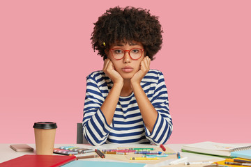 Sad dejected female architect feels unhappy as has no inspiration for creating masterpiece, sits at desktop, surrounded with crayons, notebook and disposable cup of coffee, holds chin isolated on pink