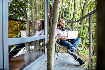 Poortrait of a handsome man working with laptop on the balcony of the house in the beautiful green forest