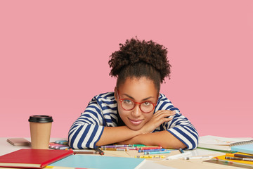 Pretty professional dark skinned designer leans on hands at workplace, looks delightfully at camera...